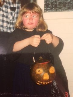 A small blonde girl dressed as a witch. Her face is painted red and she is holding a turnip lantern
