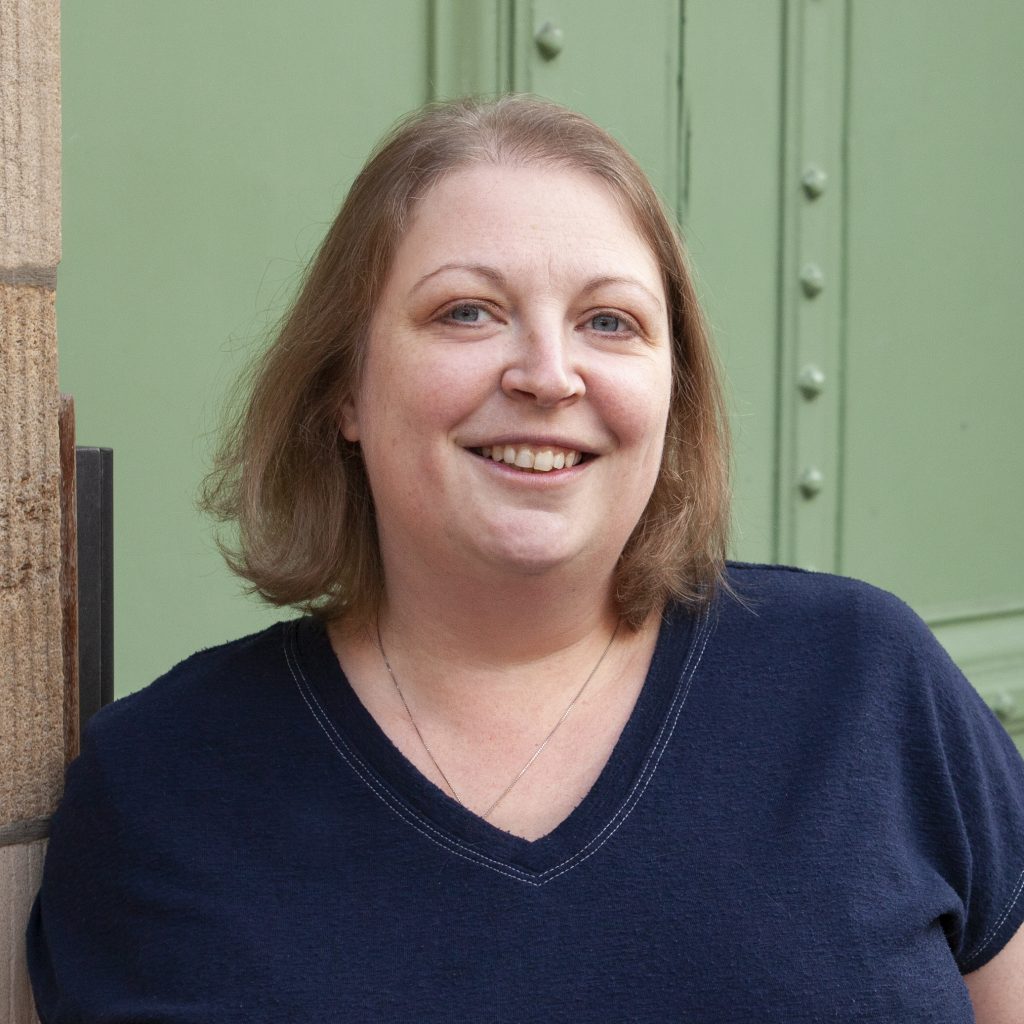 Woman in blue top with blonde hair, smiling. Standing in front of green door