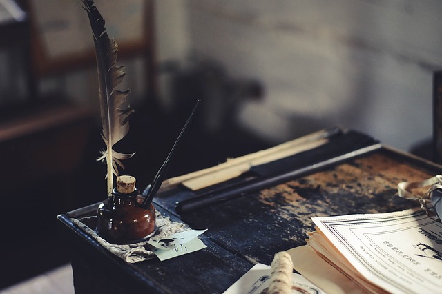 A close-up of the corner of a wooden desk. At the edge is an ink pot with a pen and a quill sticking out. A corner of a book is just visible.